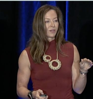 Anese Cavanaugh speaking on stage, gesturing mid-keynote under blue stage lighting