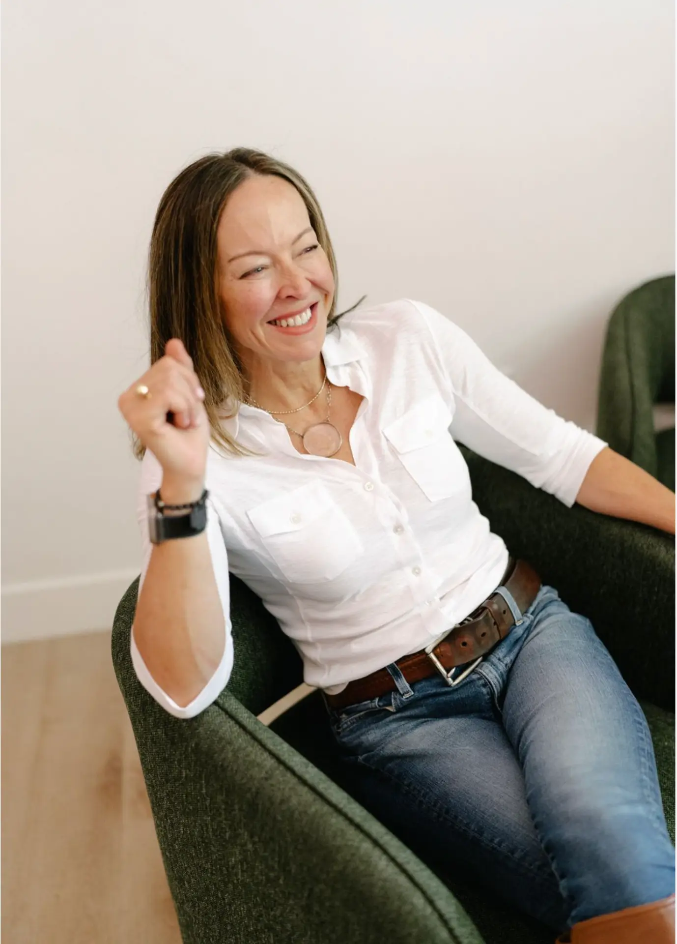 Anese Cavanaugh smiling warmly, seated in a green armchair in a white shirt and jeans