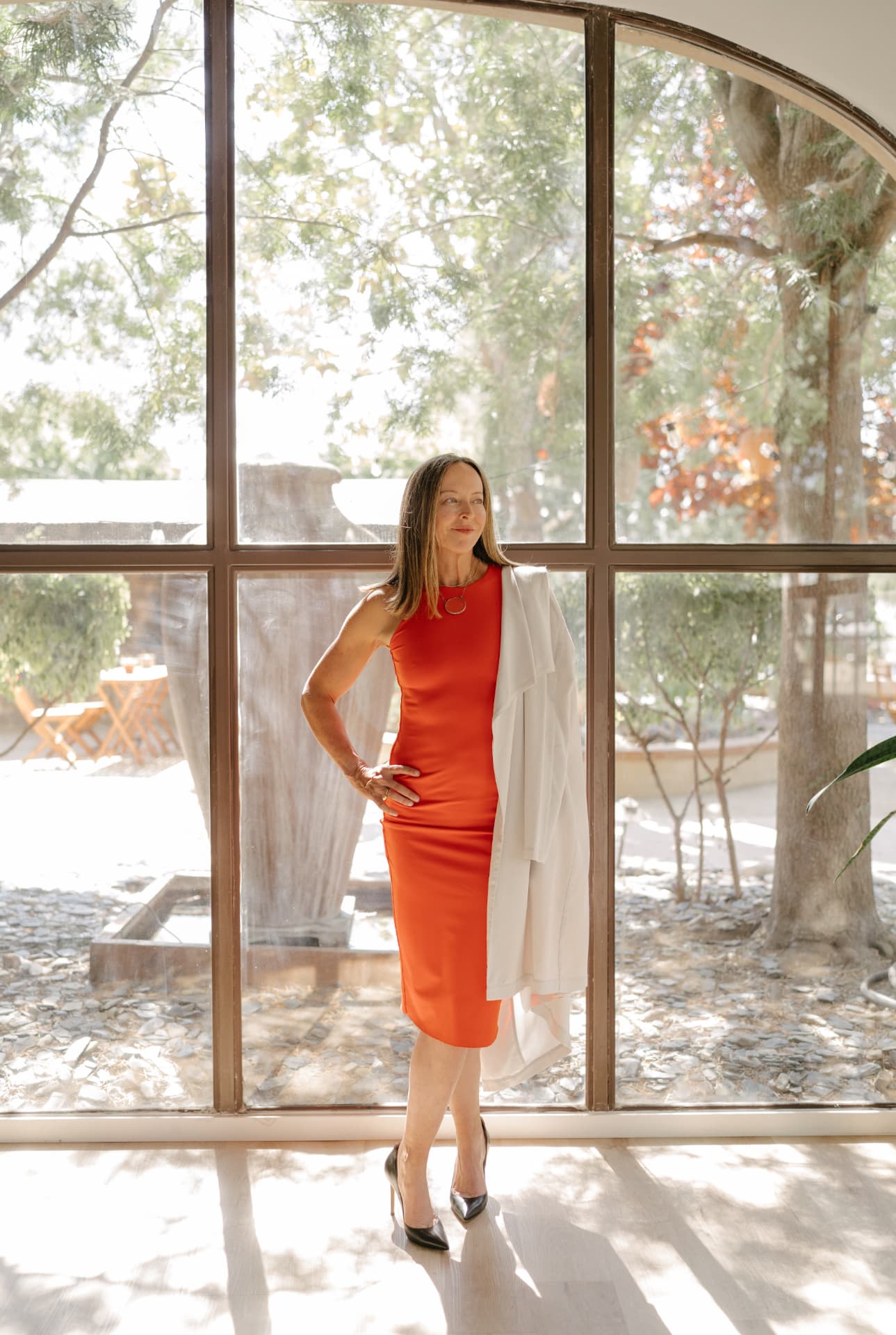 Anese Cavanaugh standing in an orange dress with a cream blazer, in front of a sunlit window