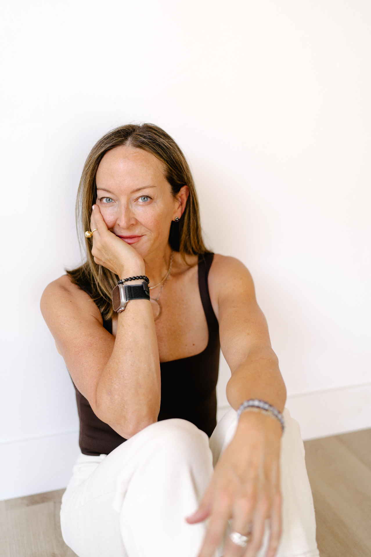 Anese seated on the floor in a brown tank and white pants, resting her chin on her hand
